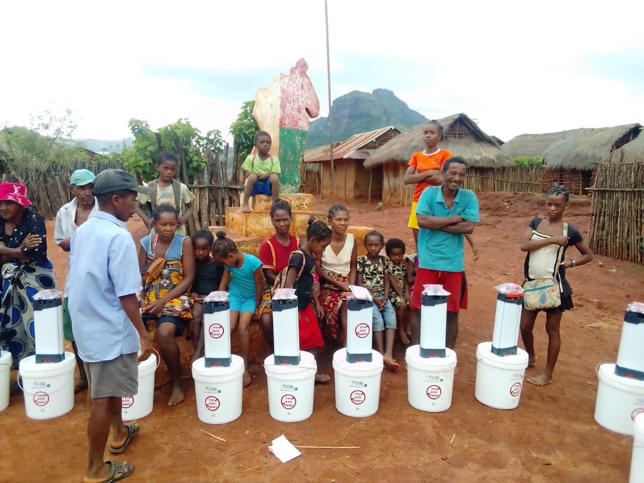 Familles réunies autour de purificateurs d’eau ORISA distribués dans un village rural à madagascar, avec enfants et habitants assis devant des habitations traditionnelles