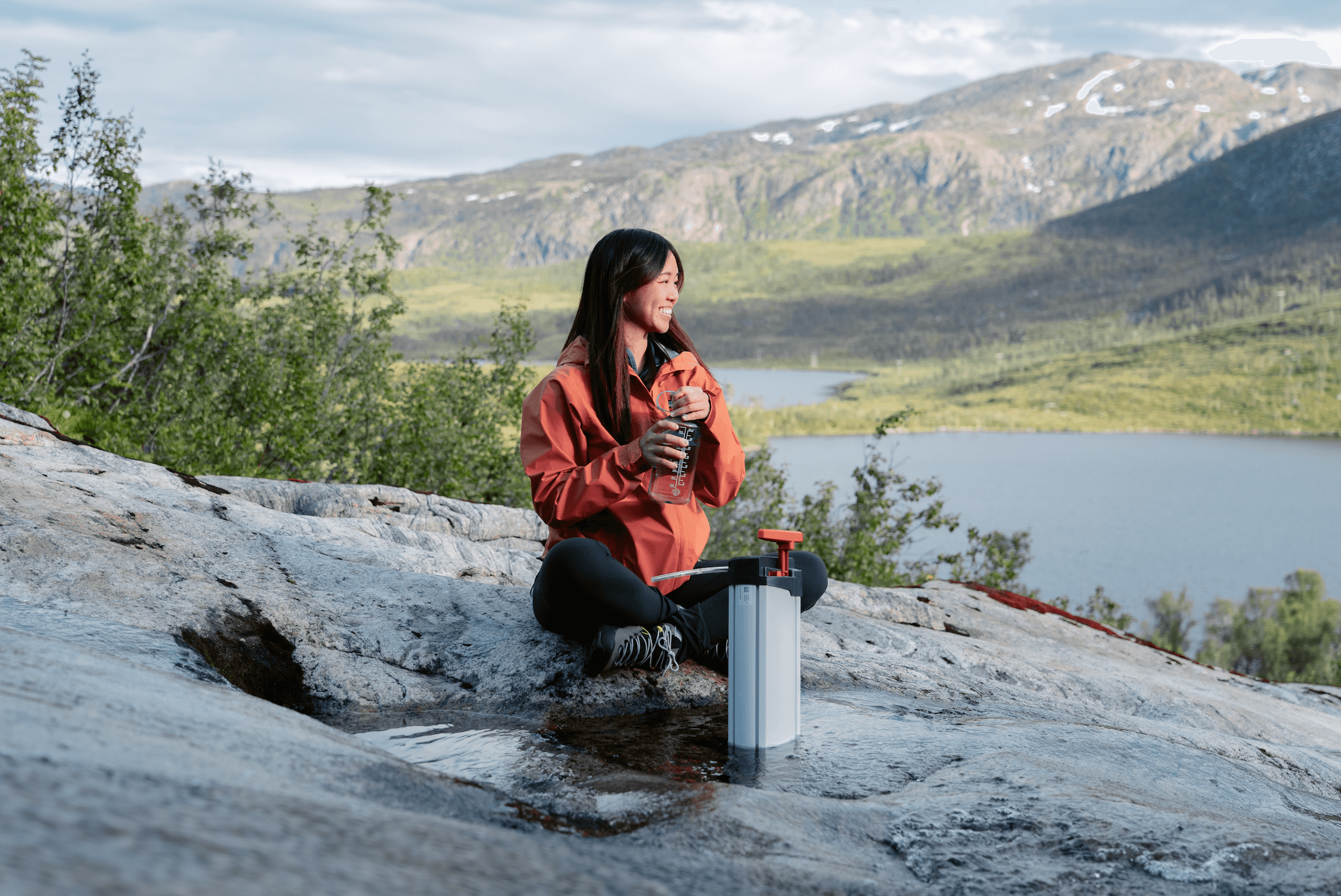 Une femme souriante après avoir utilisé le filtre à eau ORISA en pleine nature, au bord d’un ruisseau de montagne, lors d’une randonnée en autonomie